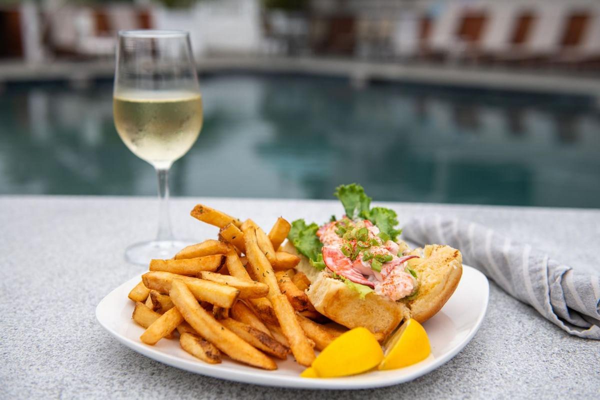 Lobster roll with fries by a pool, accompanied by a glass of white wine.
