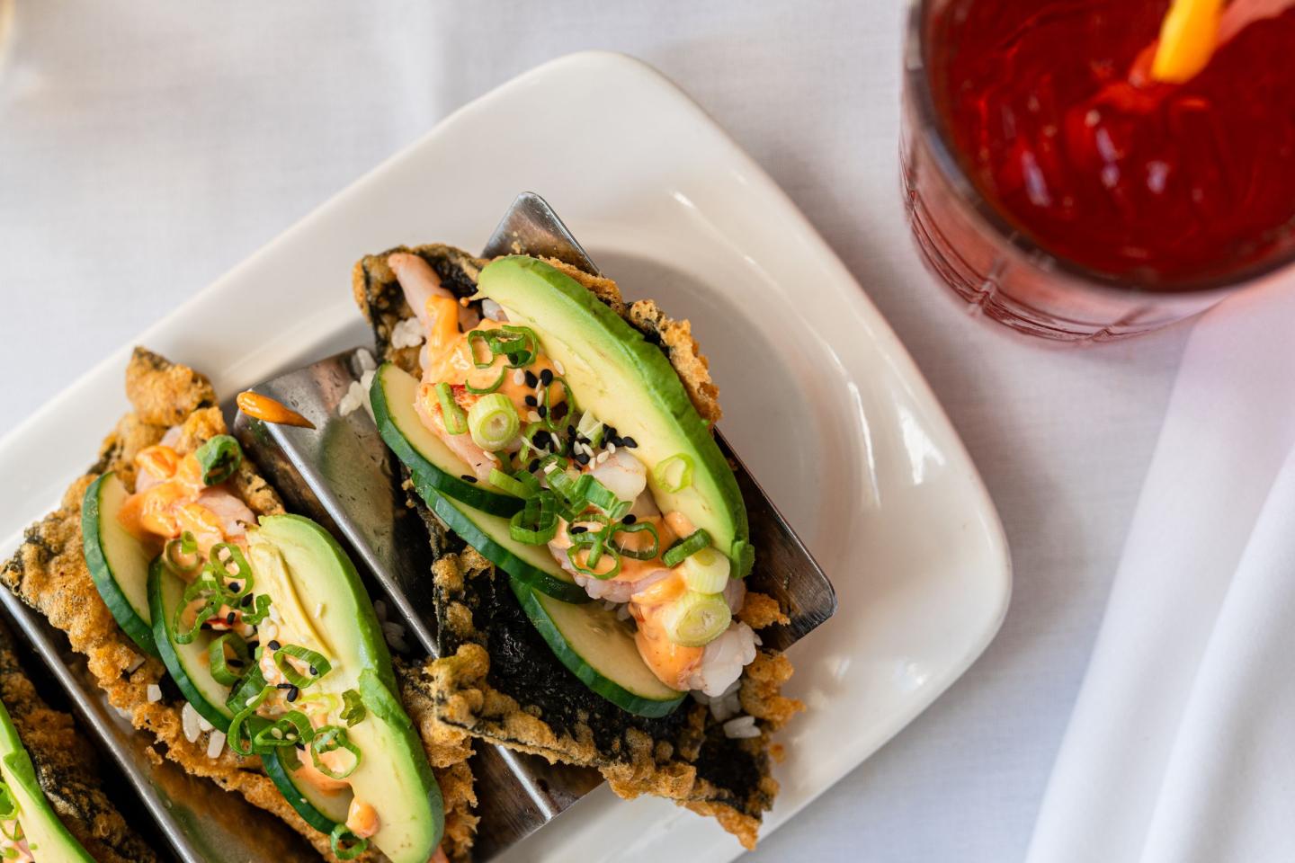 Tacos with avocado and greens next to a red drink on a white tablecloth.