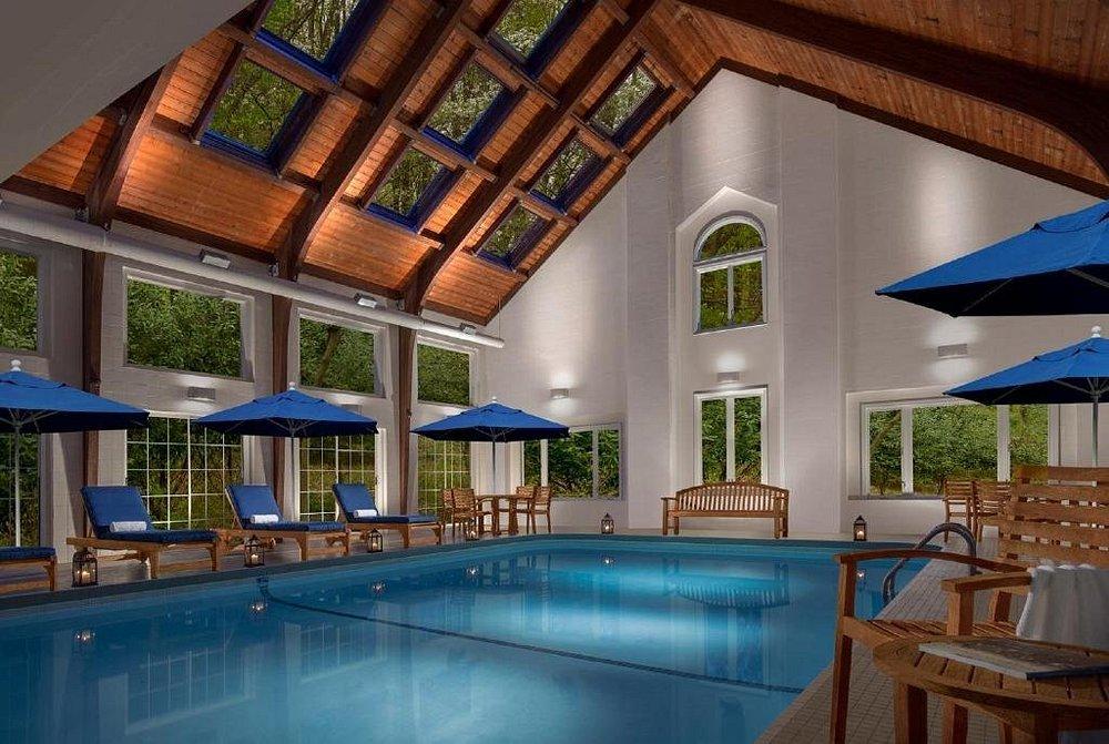 Indoor pool with blue loungers and umbrellas, wooden ceiling, large windows.