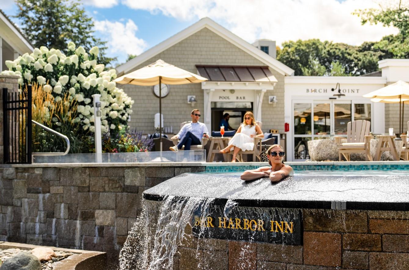 Man relaxing in a pool with a waterfall, two people sit at tables in the background.