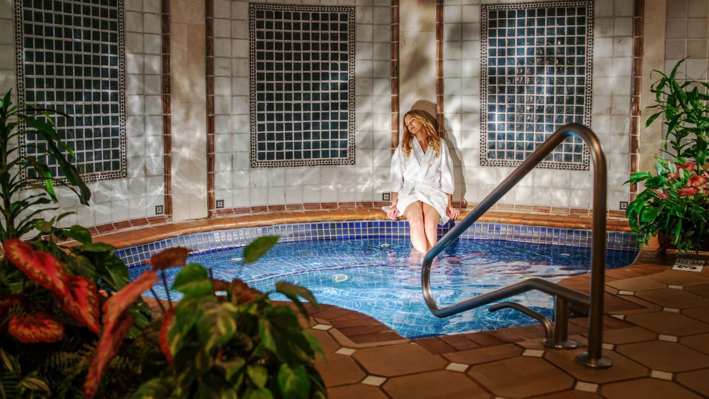 Woman in a robe sits by indoor pool surrounded by plants and tiled walls.