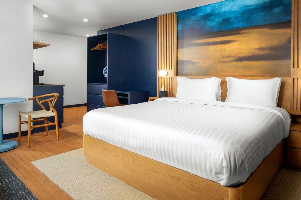 Bright hotel room with a large bed, blue accents, and a scenic wall painting.