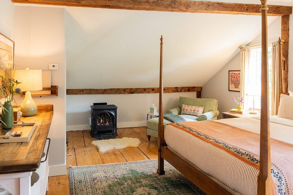 Cozy attic bedroom with wooden beams, armchair, and stove. Light from a large window.