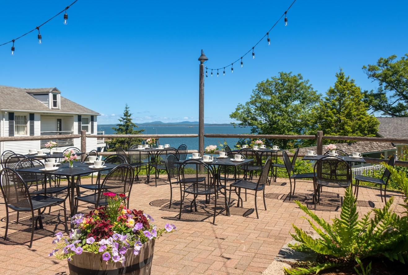 Sunny patio with tables, overlooking water, surrounded by trees and flowers.