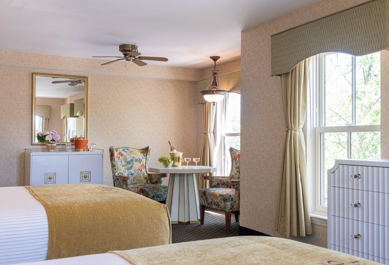 Two-bed hotel room with floral chairs, table, and large window.