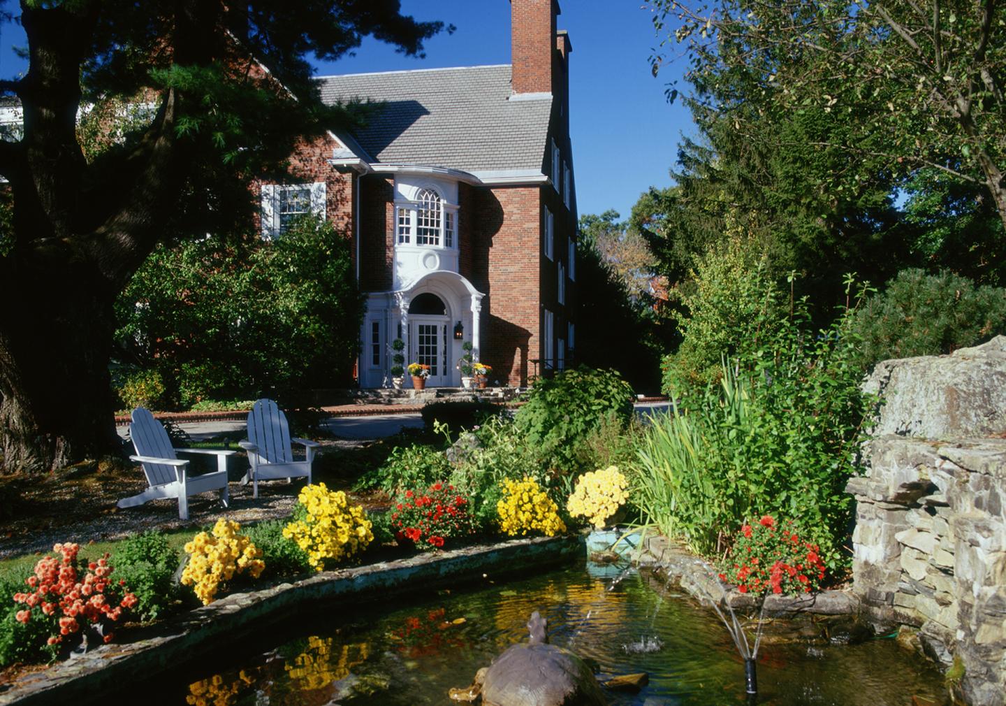 Brick house with white trim, surrounded by colorful flowers and a small pond with a fountain.