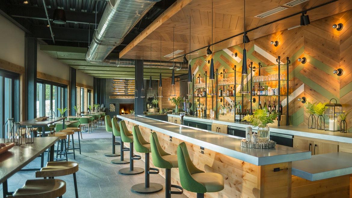 Modern bar interior with green chairs, wooden accents, and warm lighting.