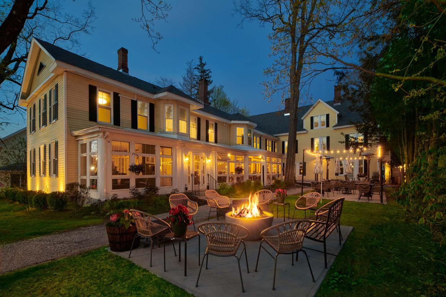 Elegant house at dusk with lit windows, fire pit, and surrounded by chairs.