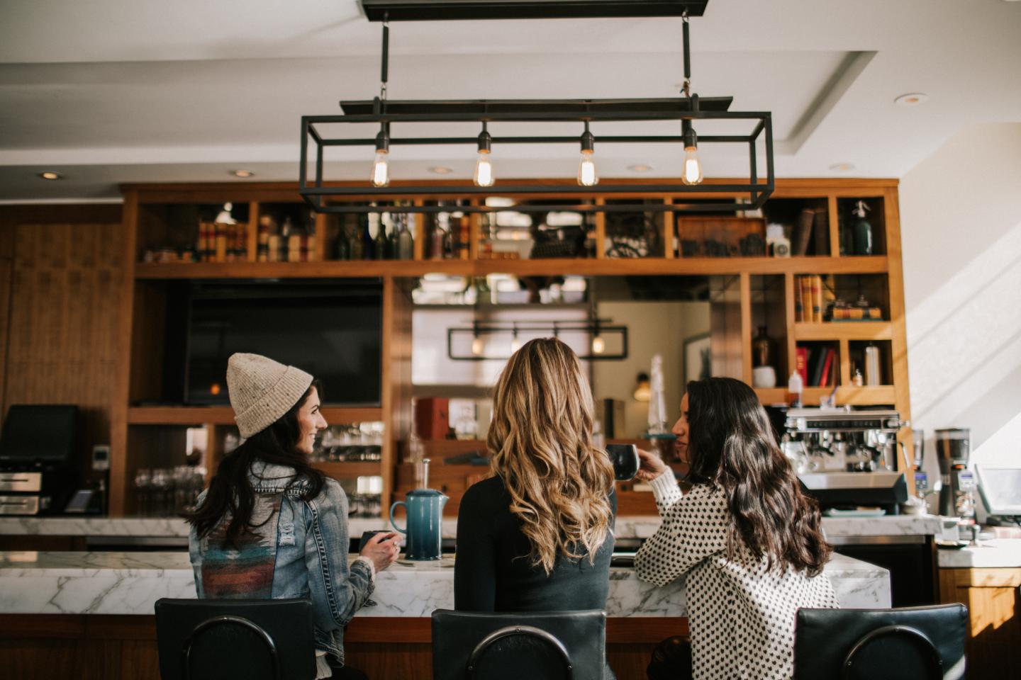 Three women seated at a café counter beneath shelves of bottles.