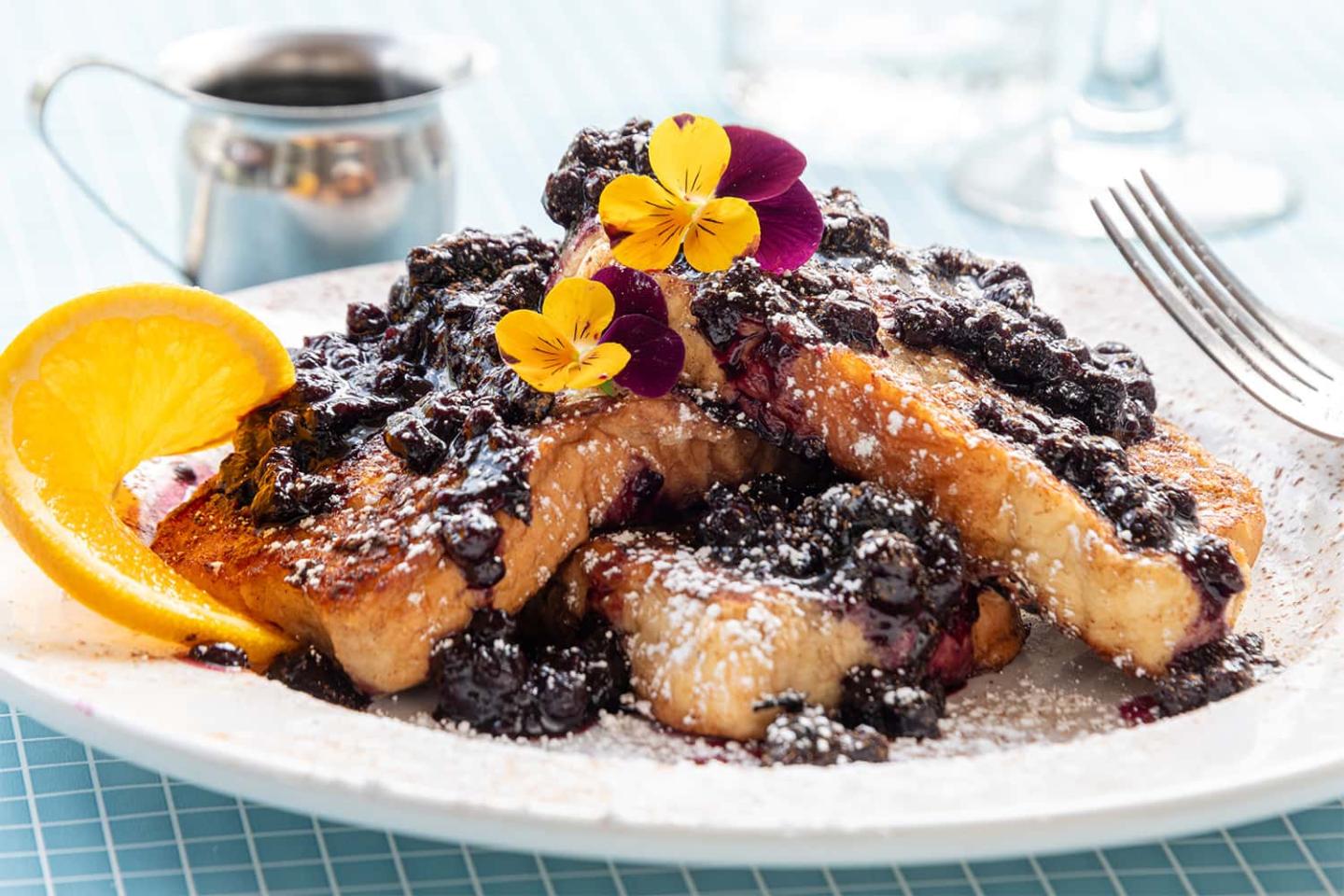 French toast with blueberries, orange slice, and yellow flowers on a white plate.