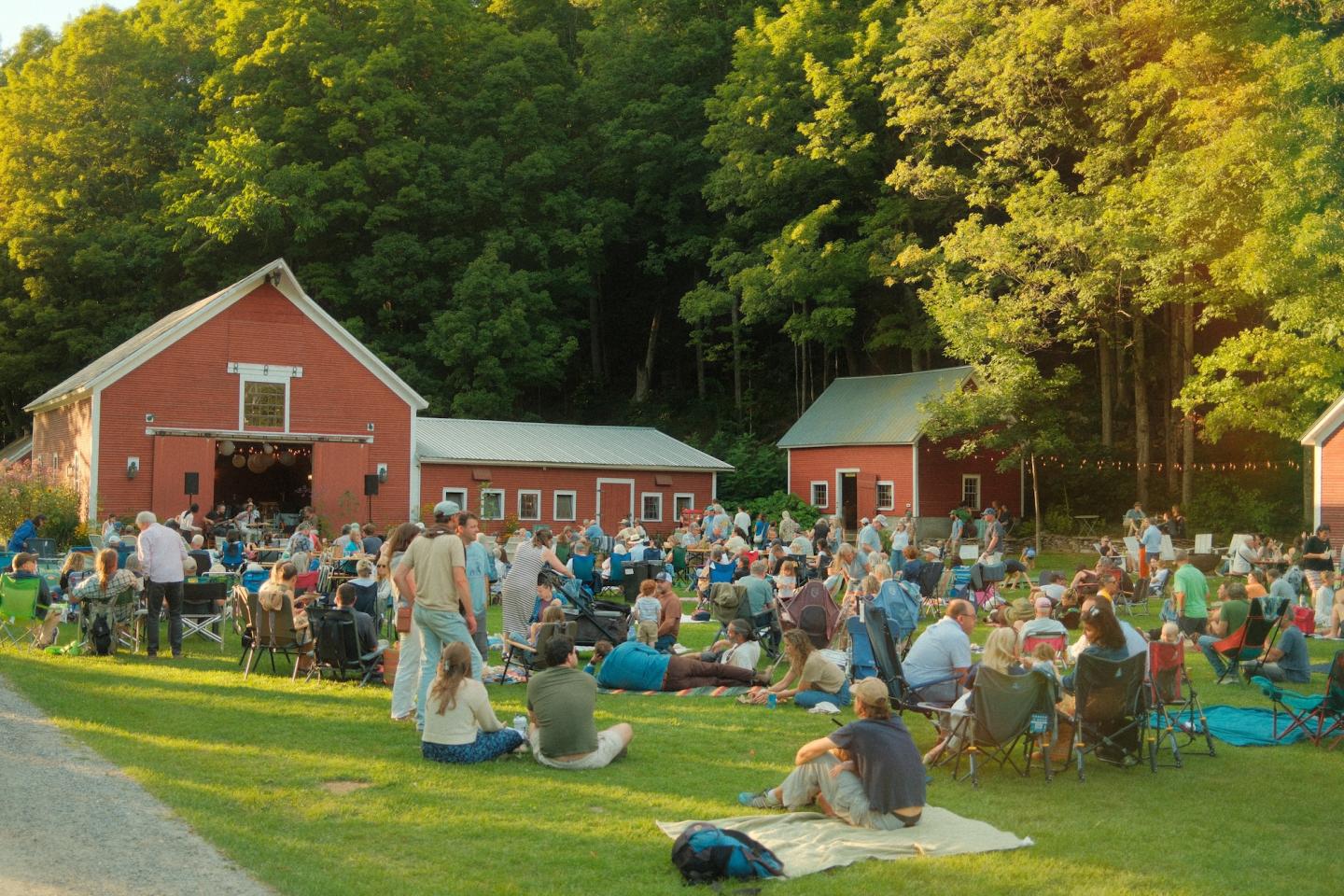 Crowd gathered outdoors near red barns, surrounded by trees.