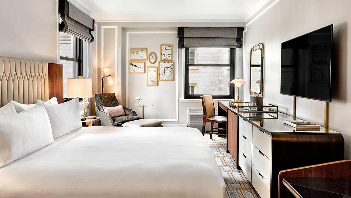 Elegant hotel room with a large bed, desk, chair, and wall-mounted TV. Neutral tones.