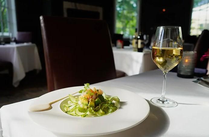 Pasta dish with green sauce and a glass of white wine on a sunlit restaurant table.