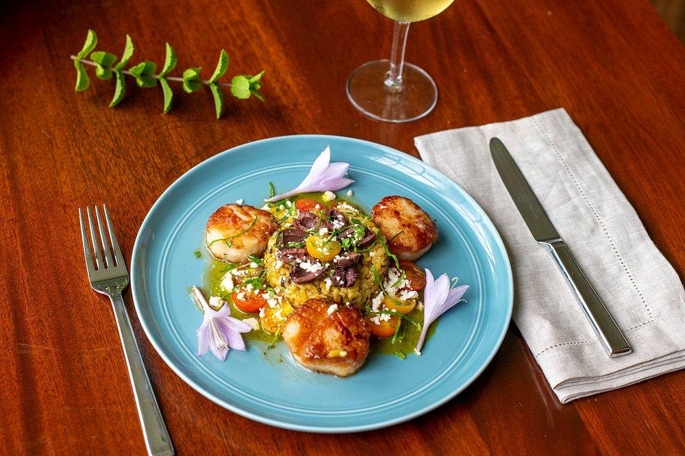 Scallops and vegetables on a blue plate, with a knife, fork, and wineglass on a wooden table.