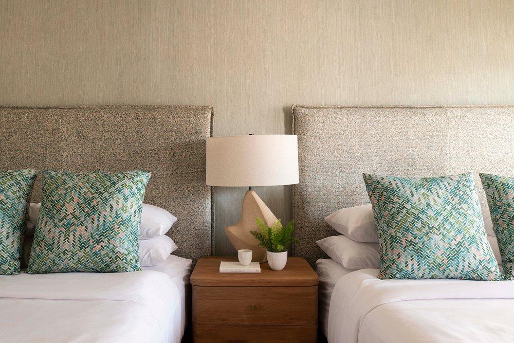 Two beds with cushions, a lamp, and a plant on a shared nightstand.
