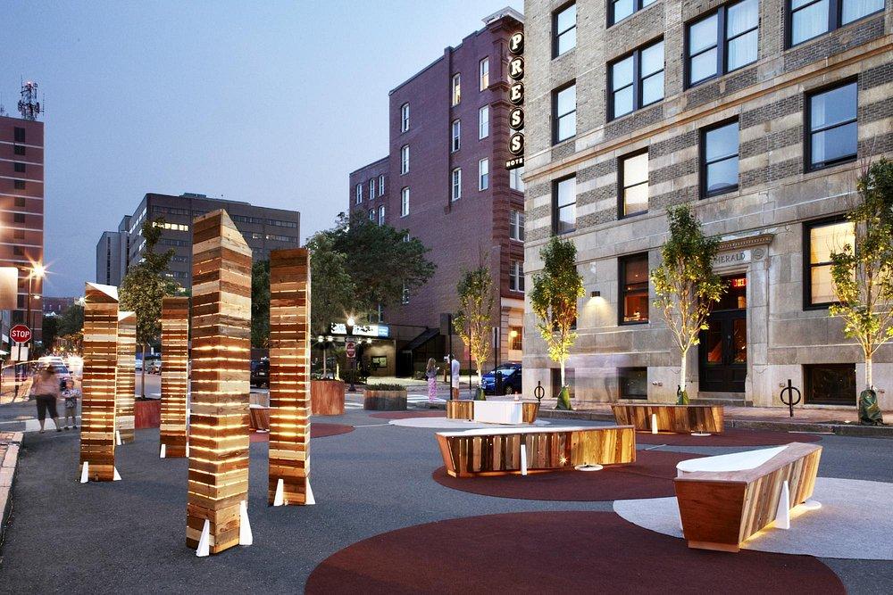 Modern plaza with wooden benches and sculptures, surrounded by brick buildings in evening light.
