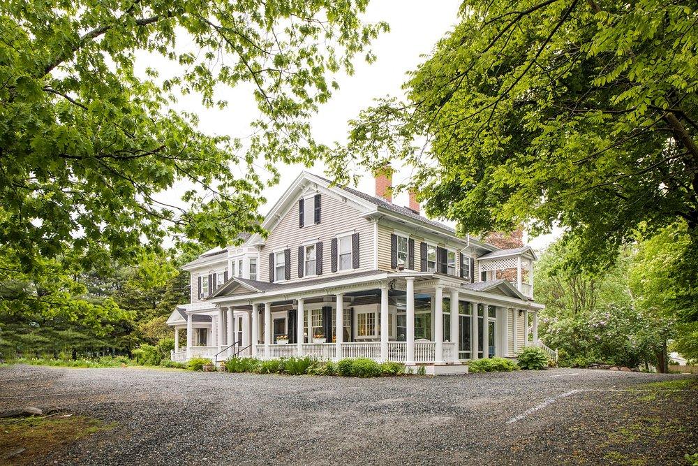 A large white house with wraparound porch surrounded by lush green trees.