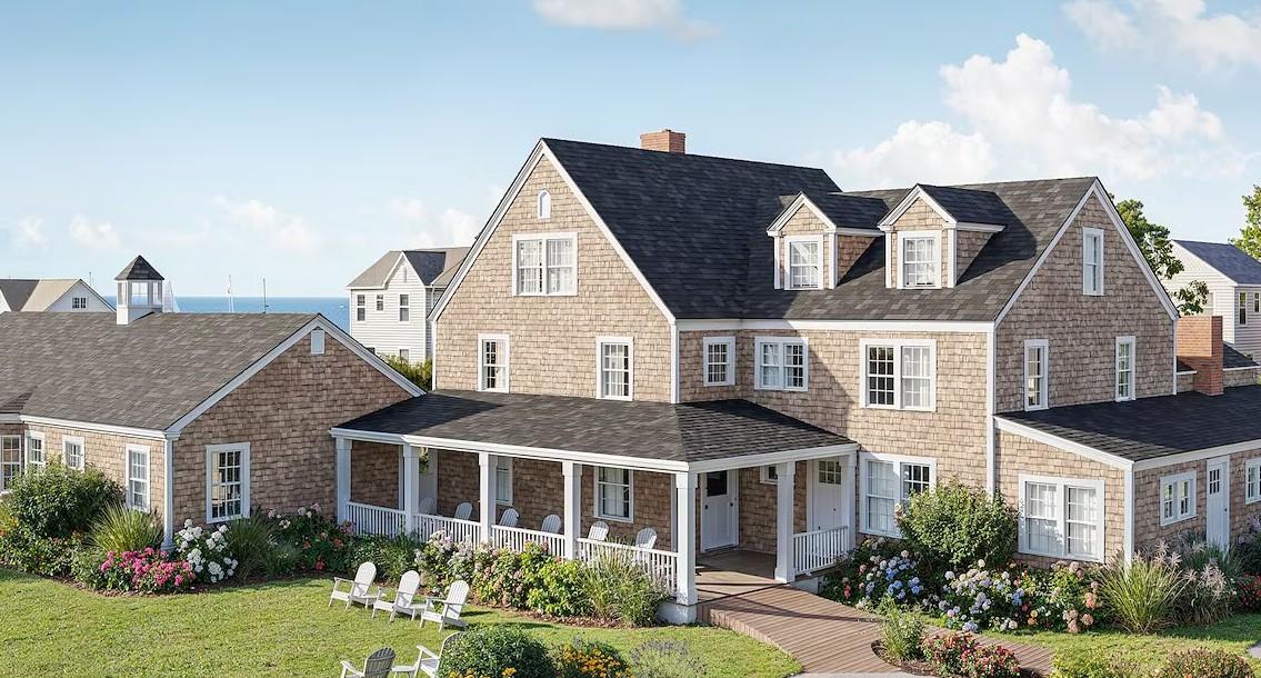 Large beige house with white trim, porch, and surrounding garden under a blue sky.