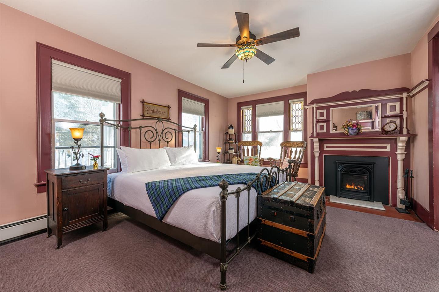 Cozy bedroom with a vintage bed, fireplace, and chest, lit by a ceiling fan lamp.