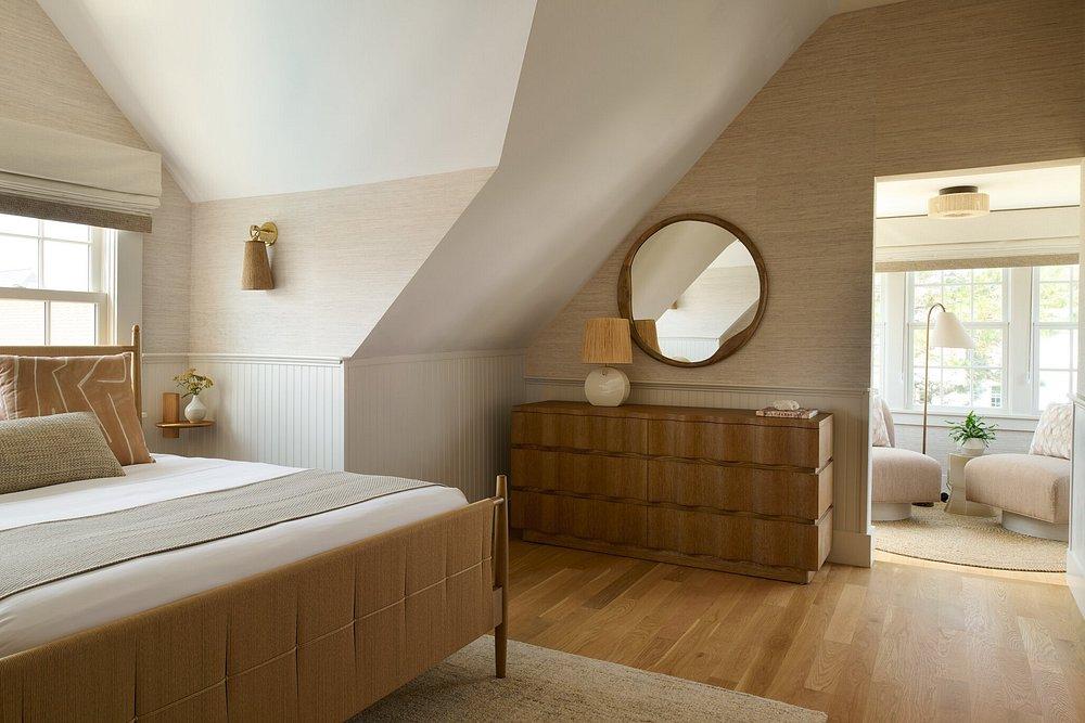 Cozy bedroom with wood accents, a round mirror, and an adjoining sunlit sitting area.