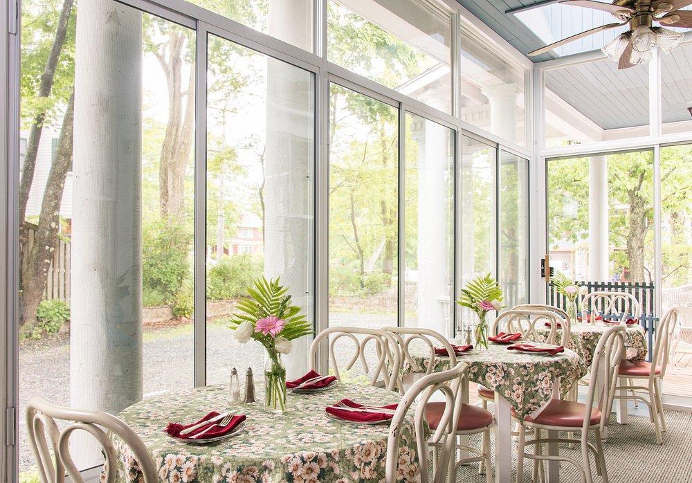 Bright dining area with floral tablecloths, red napkins, and large windows overlooking trees.
