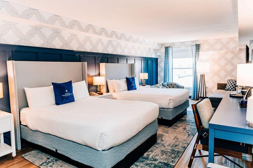 Hotel room with two double beds, blue accents, desk, and window with sheer curtains.