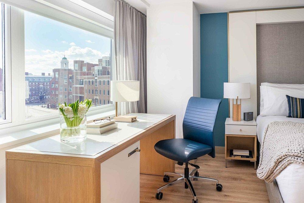 Modern hotel room with a desk, blue chair, and city view through a large window.