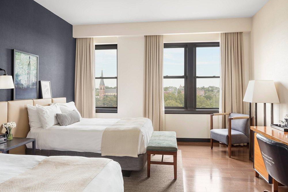 Bright hotel room with two beds, a chair, desk, and large windows.