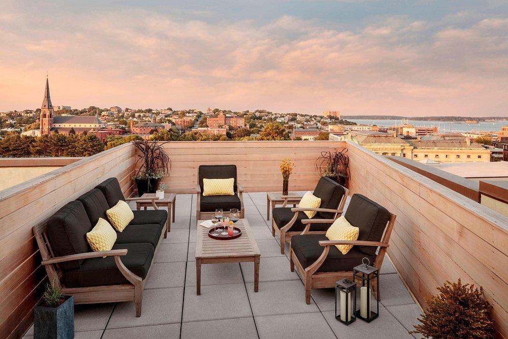 Rooftop patio with cushioned seating, wooden furniture, and sunset view over a town.