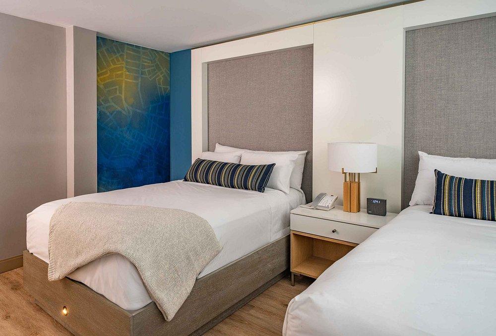 Two neatly made beds in a modern hotel room, with a lamp on the bedside table.