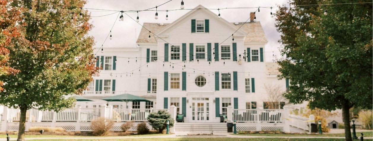 White colonial mansion with green shutters behind autumn trees.