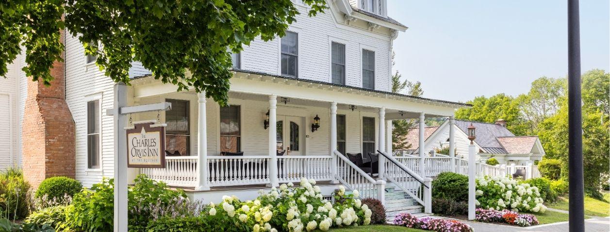White two-story house with wraparound porch, flowers, and a large tree.