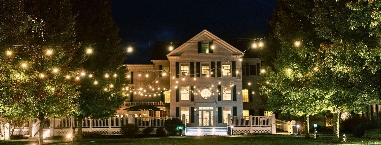 Large house warmly lit at night, surrounded by trees and string lights.