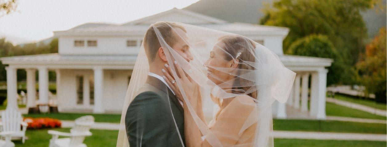 Bride and groom embrace beneath her flowing veil outside a white mansion.