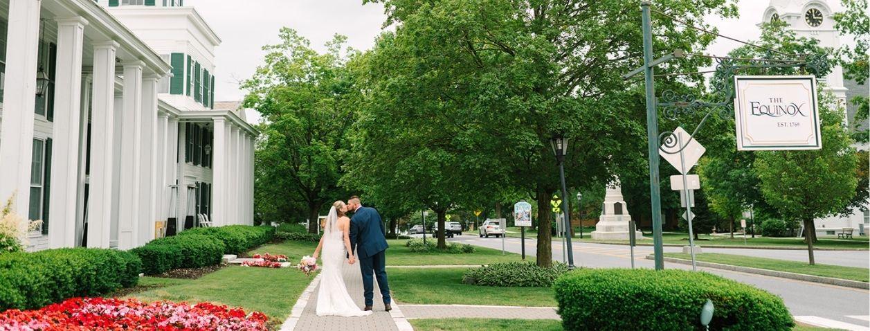 Bride and groom walking on a lawn beside red flowers and a white-columned building