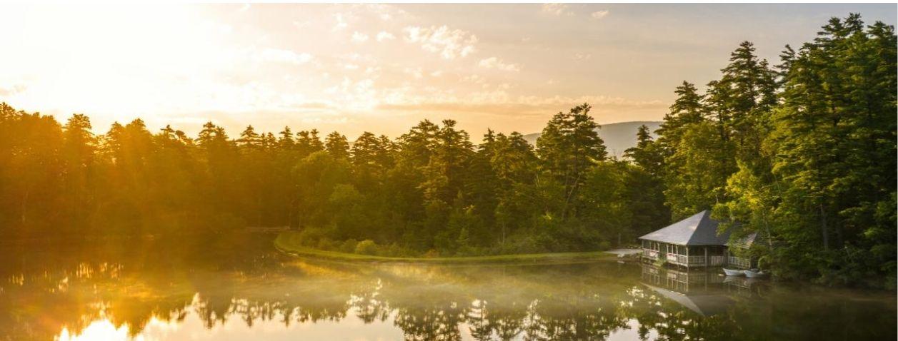 Sunset over a calm lake, reflecting trees and a small lakeside cabin.