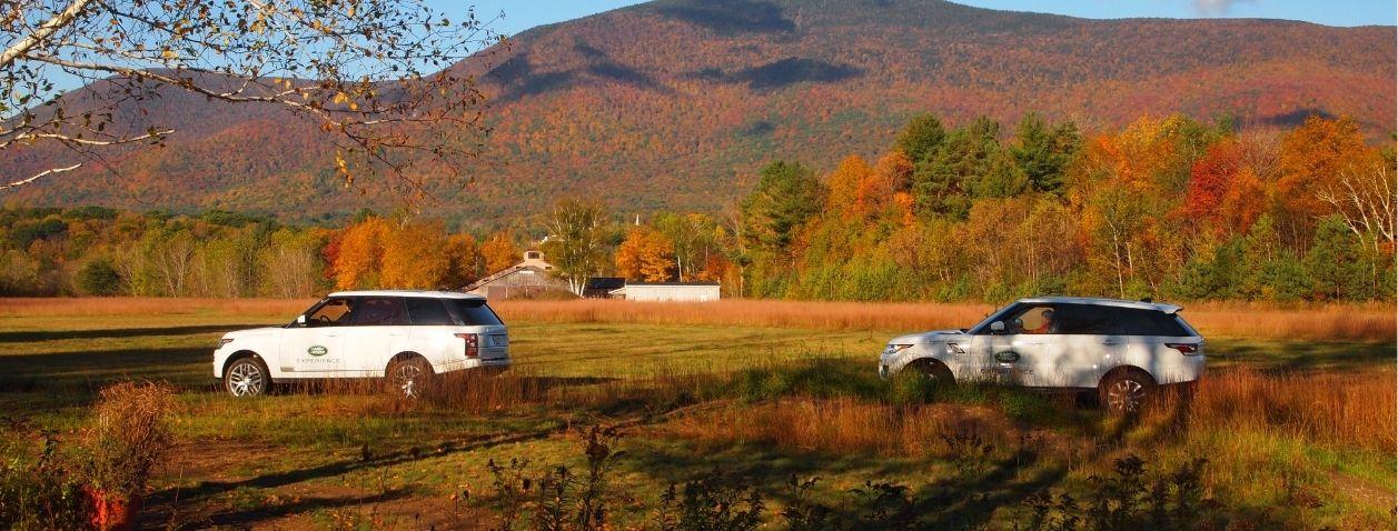 Two white cars driving on a rural road with autumn trees and mountains.