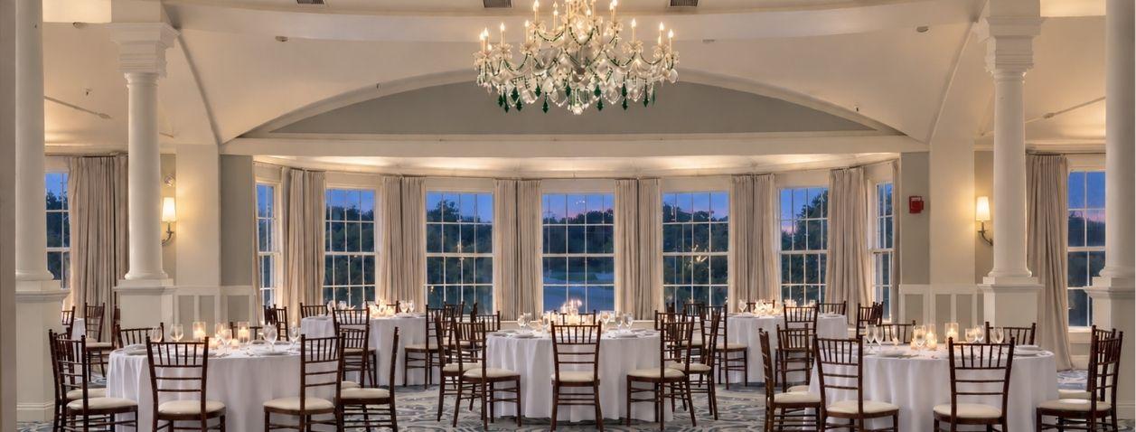 Elegant banquet hall with chandeliers, round tables, and large windows at dusk.