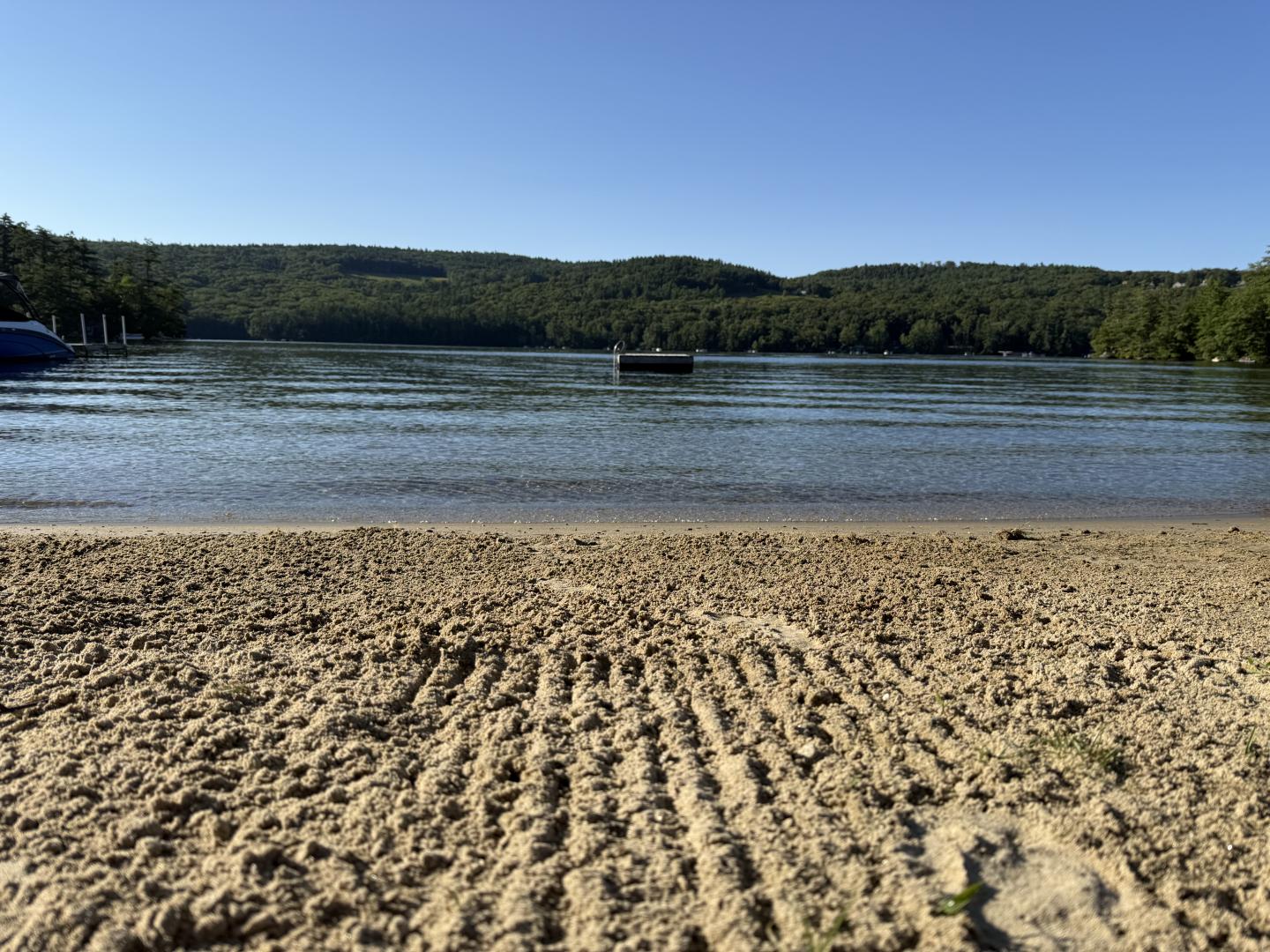 Sandy lakeshore with rippled patterns, calm water, and forested hills under blue sky.