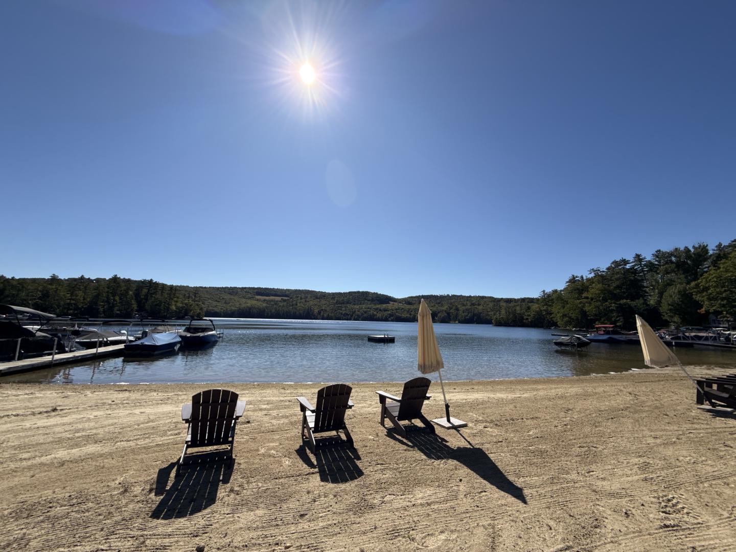 Sunny lakeside beach with chairs, umbrellas, and calm water surrounded by trees
