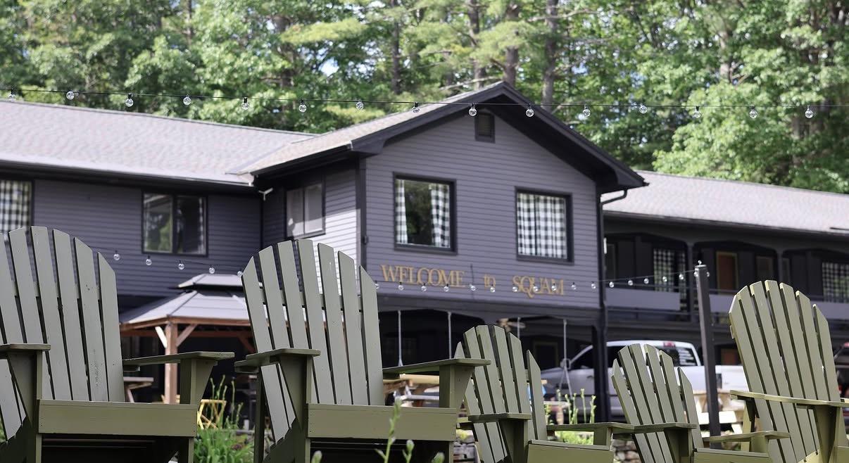 Gray lakeside cabin with Adirondack chairs on a deck beneath tall trees.