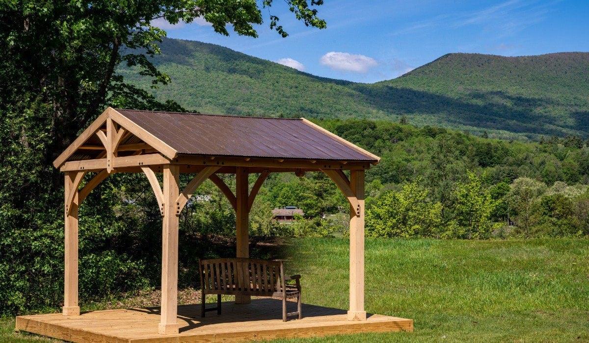 Wooden gazebo with a bench in a grassy field beneath green mountains