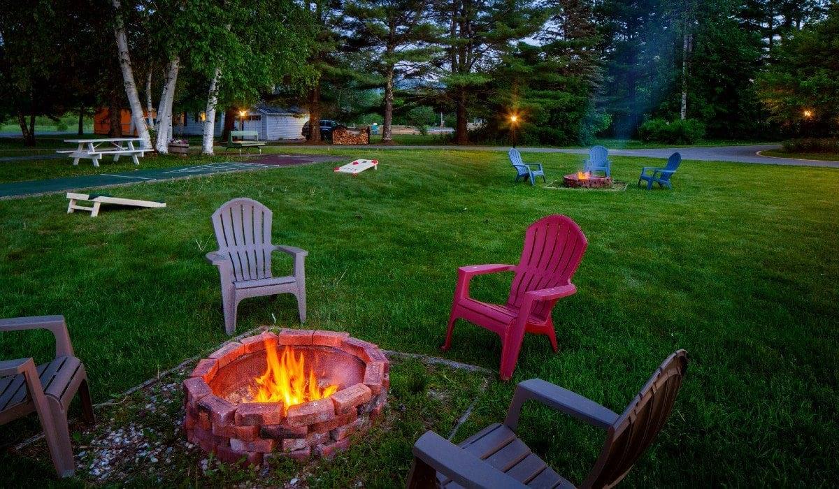 Adirondack chairs around a glowing fire pit on a grassy lawn at dusk.