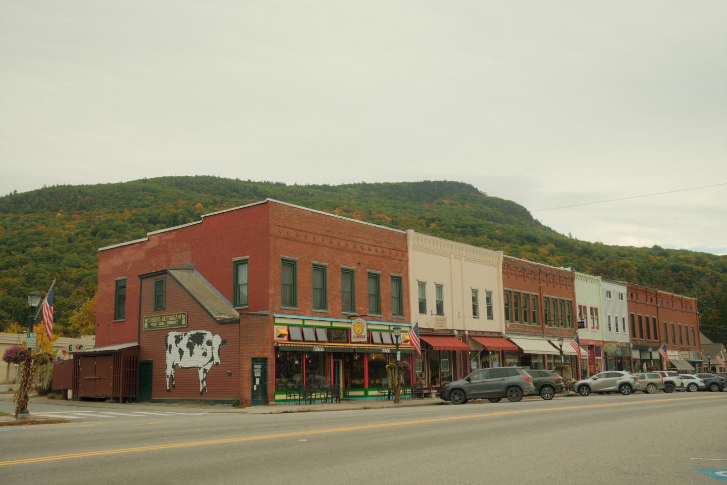 Historic brick storefronts along a quiet small-town main street beneath green hills.