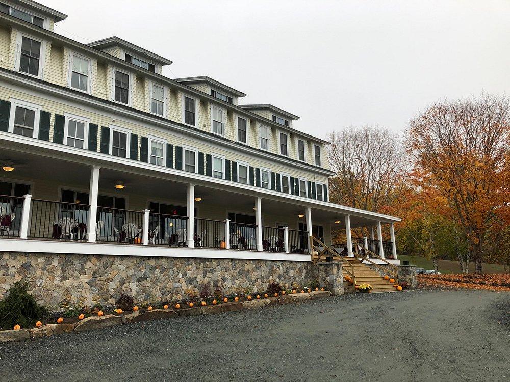 Large white inn with a wraparound porch beside autumn trees.