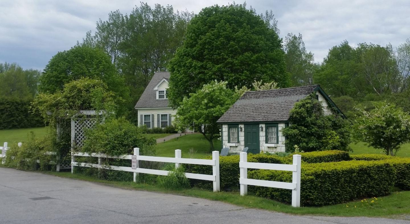 Country cottage with white fence, hedges, and large trees under a cloudy sky