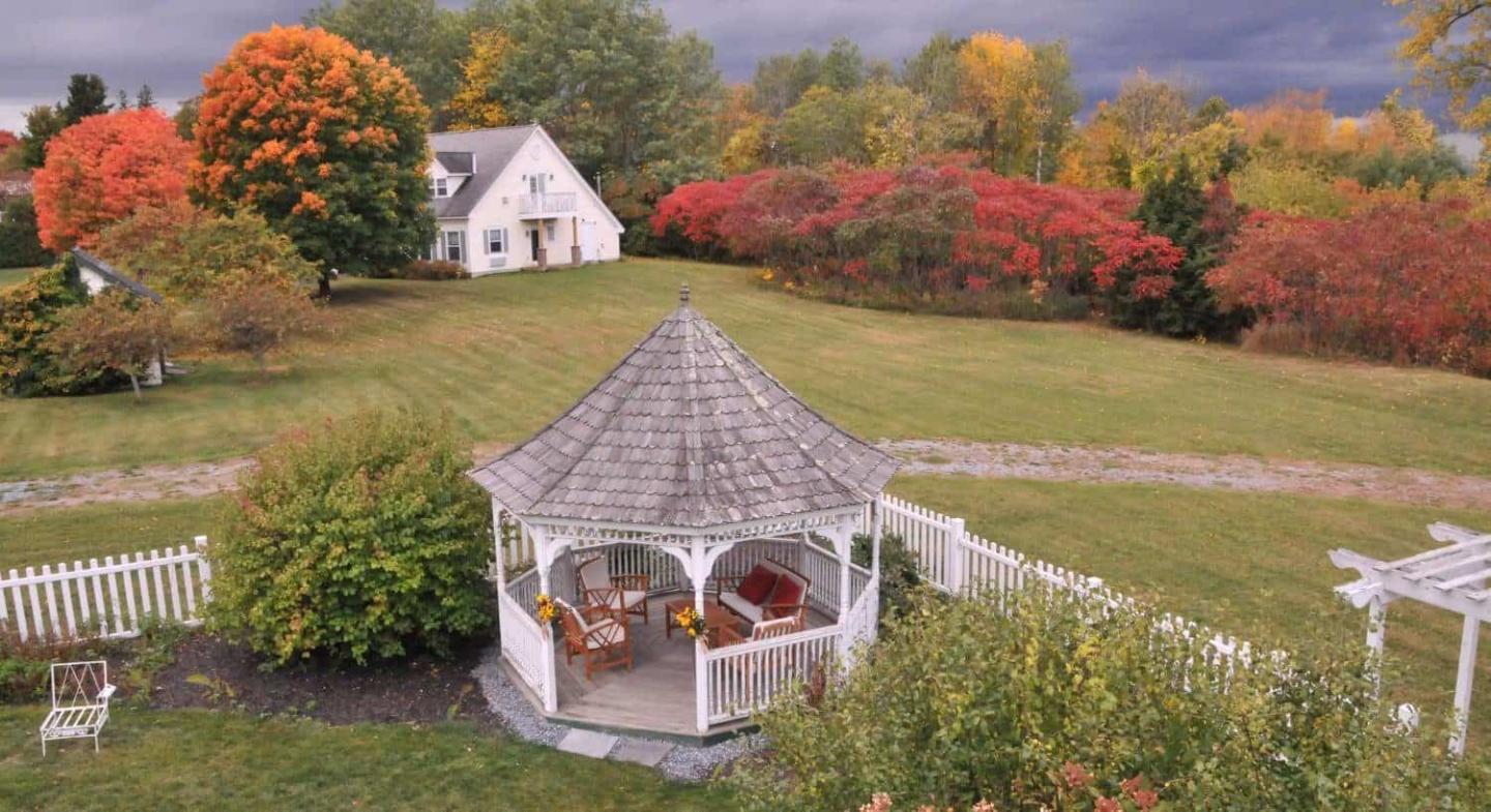 White gazebo in a flower garden, with a country house and autumn trees behind.