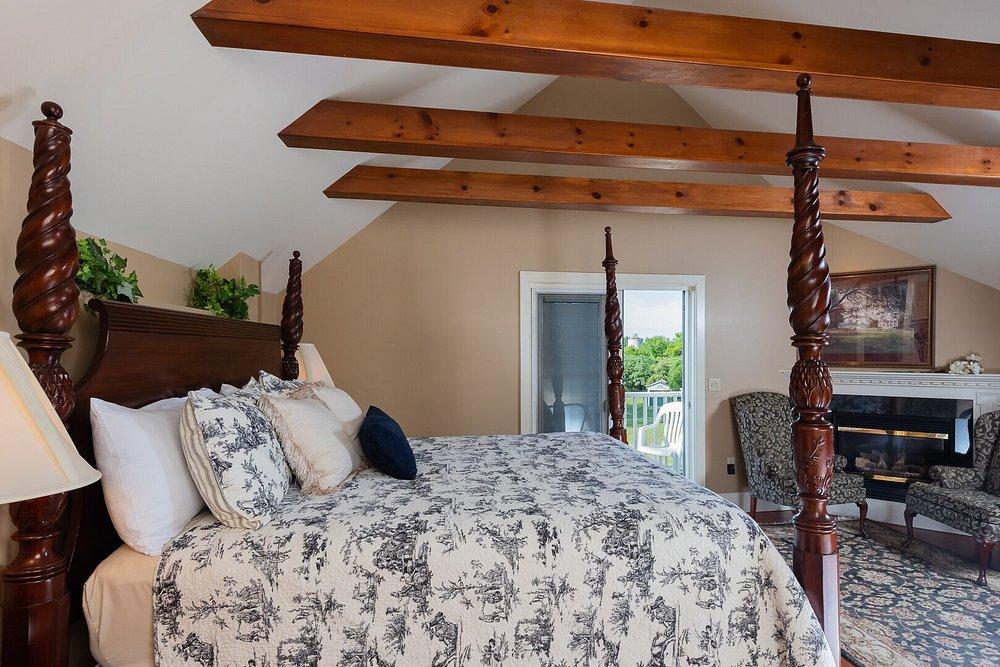Rustic bedroom with a patterned bedspread, exposed beams, and a view into a kitchen.