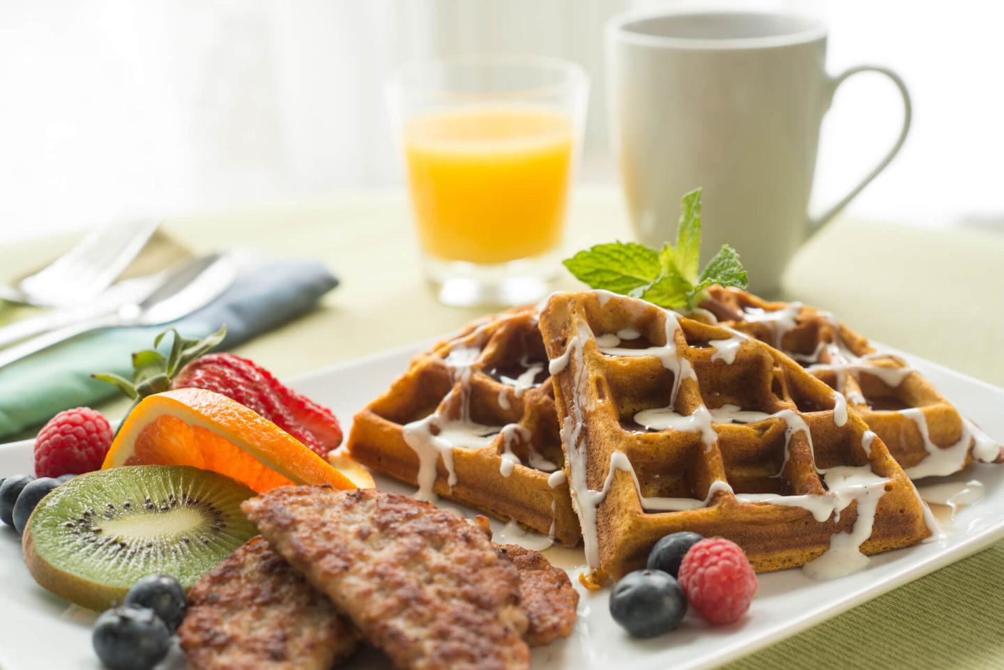 A plate with pumpkins waffles drizzled with real maple syrup and vanilla bean cream with sausage patty, fresh fruit, coffee, and orange juice.