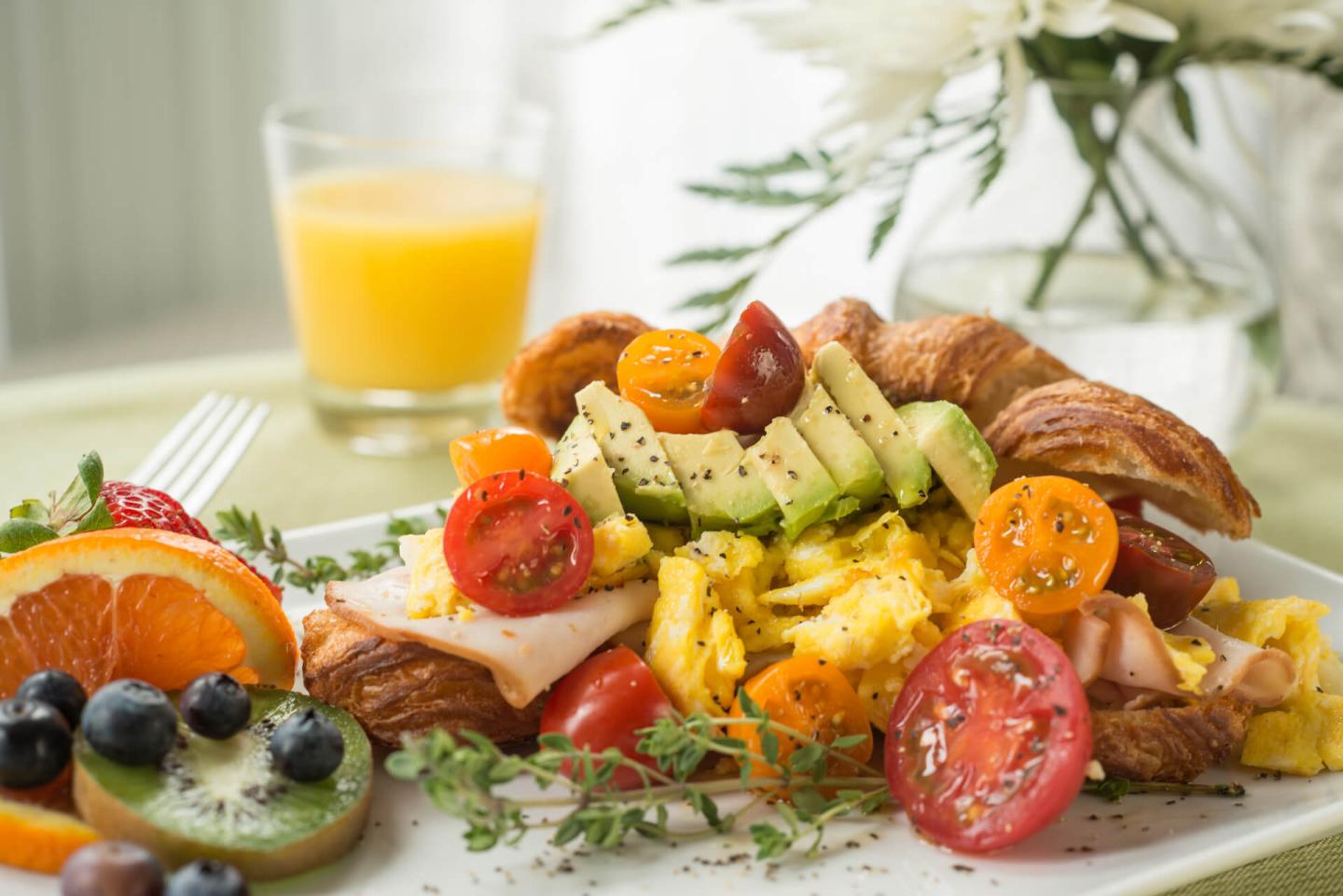 Toasted croissant with cheesy scrambled eggs, turkey, avocado, and mini heriloom tomatoes on a white plate with fresh cut fruit and a glass of orange juice in the background.